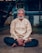 An elderly man with white hair and a bushy beard sits cross-legged on the ground, wearing a light beige tunic and dark pants. His eyes are closed, and he appears to be meditating or in deep concentration. A red tilak and white markings are on his forehead, signifying a religious or spiritual practice. The background is slightly blurred, with a building and some greenery visible.