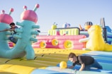 Two people are having fun in an outdoor inflatable playground with brightly colored structures and obstacles. One person is climbing a large inflatable shape resembling a creature, while another is crawling with a yellow ball in hand.