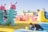 Two people are having fun in an outdoor inflatable playground with brightly colored structures and obstacles. One person is climbing a large inflatable shape resembling a creature, while another is crawling with a yellow ball in hand.