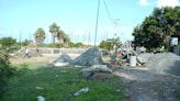 A construction site with piles of gravel and sand, surrounded by greenery and palm trees. Concrete foundations and steel rods are visible, with a cement mixer in operation. Workers can be seen in the background carrying materials.
