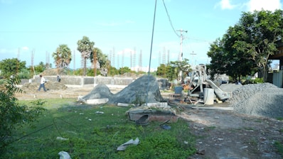 A construction site with piles of gravel and sand, surrounded by greenery and palm trees. Concrete foundations and steel rods are visible, with a cement mixer in operation. Workers can be seen in the background carrying materials.