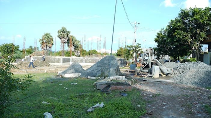 A construction site with piles of gravel and sand, surrounded by greenery and palm trees. Concrete foundations and steel rods are visible, with a cement mixer in operation. Workers can be seen in the background carrying materials.