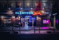 A barbershop storefront with vibrant neon signage and a modern industrial exterior. The shop is called 'Mr. Brothers Cut Club' and features a mix of illuminated blue, red, and purple signs, creating a lively and urban atmosphere. Outside, bicycles are parked, and a person sits casually on a bench.