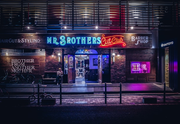 A barbershop storefront with vibrant neon signage and a modern industrial exterior. The shop is called 'Mr. Brothers Cut Club' and features a mix of illuminated blue, red, and purple signs, creating a lively and urban atmosphere. Outside, bicycles are parked, and a person sits casually on a bench.