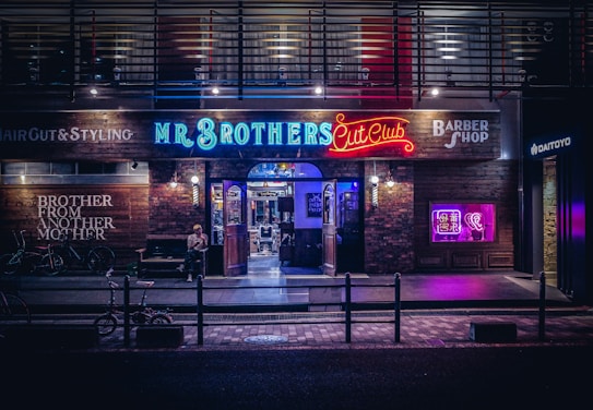 A barbershop storefront with vibrant neon signage and a modern industrial exterior. The shop is called 'Mr. Brothers Cut Club' and features a mix of illuminated blue, red, and purple signs, creating a lively and urban atmosphere. Outside, bicycles are parked, and a person sits casually on a bench.