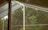 A view through a glass-covered patio or veranda, showcasing lush green trees outside. The structure features wooden and white metal elements, creating a contrast with the natural greenery.