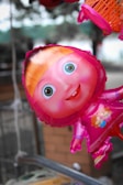 A colorful, cartoon character-shaped balloon with a large, smiling face is hanging outdoors. The character has large, vibrant eyes and orange hair, and is wearing a pink outfit. The background is slightly blurred, indicating an outdoor setting.