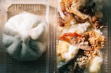 A clear plastic container holds a Chinese steamed bun on the left and a portion of rice topped with various ingredients, including cooked onions, roasted meat, and fried shallots on the right.