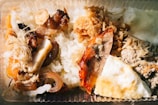 A close-up view of a meal served in a clear plastic container. The dish includes white rice and various toppings such as grilled meat, roasted vegetables, shredded dried toppings, and a portion of fried egg.