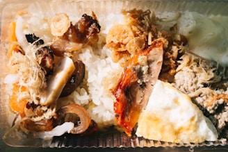 A close-up view of a meal served in a clear plastic container. The dish includes white rice and various toppings such as grilled meat, roasted vegetables, shredded dried toppings, and a portion of fried egg.