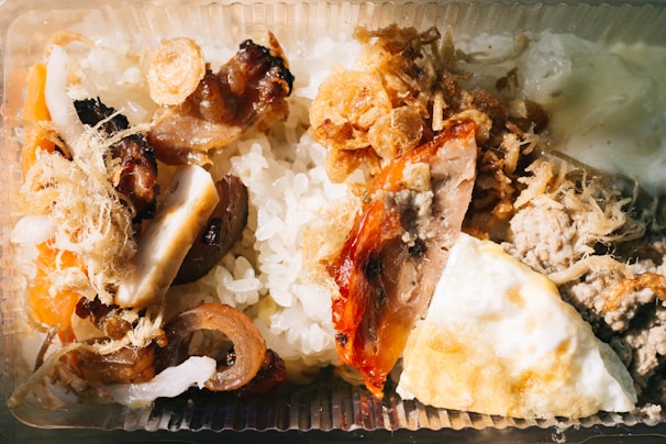 A close-up view of a meal served in a clear plastic container. The dish includes white rice and various toppings such as grilled meat, roasted vegetables, shredded dried toppings, and a portion of fried egg.