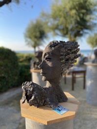 A sculpted bust is prominently displayed on a wooden pedestal. The sculpture is dark in color and appears to have an organic, textured form, resembling wind-swept hair and a serene facial expression. It is set in an outdoor environment with blurred trees and a clear sky in the background, creating a serene and natural setting. A small card with images lies on the pedestal beside the sculpture.