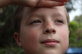 A close-up of a young child's face with eyes gazing upward. A hand is shielding the child's eyes from the light. The background is softly blurred with greenery, suggesting an outdoor setting.