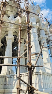 Detailed white stone architecture with intricate carvings and ornate pillars. Wooden scaffolding with ropes is in place, indicating construction or restoration work. The background shows a clear blue sky with a few clouds.