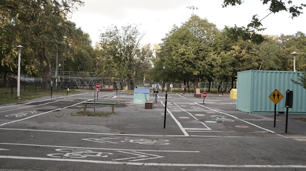 An empty park area featuring a mini traffic layout with bike paths, road signs, and small structures. There are trees lining the path and a green container on the side. The ground is marked with various road symbols and pedestrian crossings.