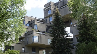A complex architectural structure composed of stacked, cubic concrete modules, each featuring large windows. The building is surrounded by lush green trees and vegetation. The sky is partially visible, showing a clear day.