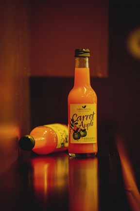 Two bottles of organic carrot and apple juice are shown, with one bottle standing upright and the other lying on its side. The setting has a warm, dim lighting that creates a cozy atmosphere.