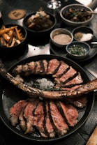 A large, cooked tomahawk steak sliced into pieces is presented on a black plate. The surrounding table features various side dishes, including cooked vegetables, sauces, and condiments. The atmosphere suggests a gourmet dining setting with elegant plating.