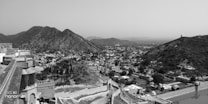 A vast landscape view of a historic fort or palace situated on a hillside overlooking a sprawling town or city. The architecture is a mix of stone walls and traditional design elements. Surrounding the town are rugged hills, indicating a region rich in history and natural beauty, all captured in black and white, adding a timeless feel to the scene.
