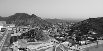 A vast landscape view of a historic fort or palace situated on a hillside overlooking a sprawling town or city. The architecture is a mix of stone walls and traditional design elements. Surrounding the town are rugged hills, indicating a region rich in history and natural beauty, all captured in black and white, adding a timeless feel to the scene.