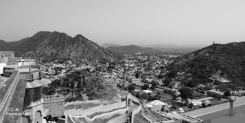 A vast landscape view of a historic fort or palace situated on a hillside overlooking a sprawling town or city. The architecture is a mix of stone walls and traditional design elements. Surrounding the town are rugged hills, indicating a region rich in history and natural beauty, all captured in black and white, adding a timeless feel to the scene.
