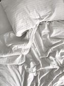 A neatly arranged bed with white sheets and a pillow, featuring slightly crumpled fabric creating light and shadow play.