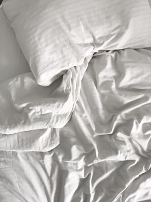 A neatly arranged bed with white sheets and a pillow, featuring slightly crumpled fabric creating light and shadow play.