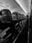 A black and white view of an airplane cabin with empty seats in rows along the aisle. Overhead compartments and lighting are visible, and the interior has a sleek, modern design. The atmosphere is calm and orderly, with symmetrical lines leading towards the back of the plane.