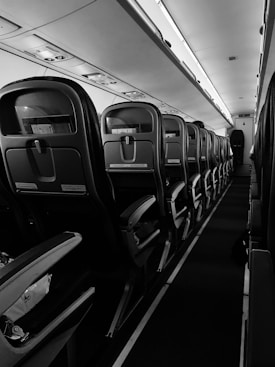 A black and white view of an airplane cabin with empty seats in rows along the aisle. Overhead compartments and lighting are visible, and the interior has a sleek, modern design. The atmosphere is calm and orderly, with symmetrical lines leading towards the back of the plane.