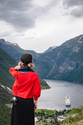 A person wearing a black beret, red shirt, and black pants is standing on a hill overlooking a fjord. A camera hangs from their side. In the distance, a cruise ship travels through the water surrounded by steep, green mountains under a cloudy sky.