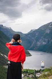 A person wearing a black beret, red shirt, and black pants is standing on a hill overlooking a fjord. A camera hangs from their side. In the distance, a cruise ship travels through the water surrounded by steep, green mountains under a cloudy sky.