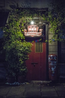 A red door surrounded by lush green plants is illuminated by a warm light above, giving it a cozy appearance. A hanging sign with 'The Postcard Shop' is visible, and a vintage mailbox covered in stickers stands to the right of the door.
