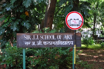 A wooden sign with white text reads 'SIR J.J. SCHOOL OF ART' alongside its Hindi translation. The sign is mounted on green metal posts amidst lush greenery, including various large-leafed plants. To the right, a circular red and white sign with an illustration of a fence and a notice in Hindi is visible.