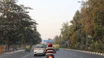 A road scene with a person wearing a red helmet riding a motorcycle. The road is flanked by trees on both sides, with several cars and auto-rickshaws moving along. The sky is clear, suggesting a calm day.