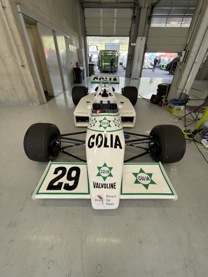 A white race car with the number 29 prominently displayed on the front wing is parked inside a garage. The car features branding and logos for Golia and Valvoline, among others. The garage is spacious with concrete walls, and a partially open roller door reveals a green vehicle outside. Various equipment, including tires and cables, are visible in the garage.