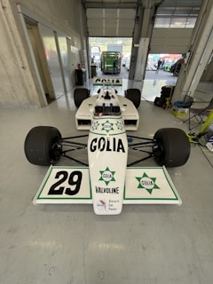 A white race car with the number 29 prominently displayed on the front wing is parked inside a garage. The car features branding and logos for Golia and Valvoline, among others. The garage is spacious with concrete walls, and a partially open roller door reveals a green vehicle outside. Various equipment, including tires and cables, are visible in the garage.