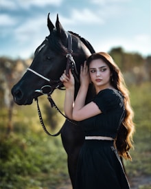 A woman with long hair is standing beside a black horse in a natural setting. She is gently holding the horse's bridle and looking to the side, both appearing calm and serene. The background features a blurred landscape with green vegetation and a soft blue sky.