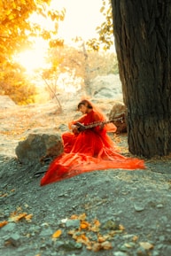 A person wearing a flowing red dress is seated on the ground near a large tree, playing a string instrument similar to a sitar. The setting is a natural outdoor environment with rocks and a sunny, autumnal background featuring warm, golden light filtering through the foliage.