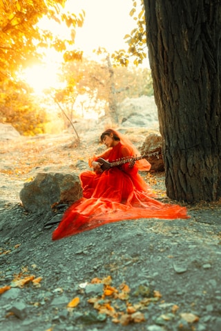 A person wearing a flowing red dress is seated on the ground near a large tree, playing a string instrument similar to a sitar. The setting is a natural outdoor environment with rocks and a sunny, autumnal background featuring warm, golden light filtering through the foliage.