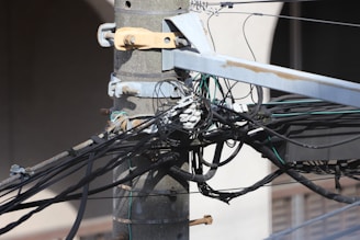 A close-up of a utility pole with numerous tangled cables and wires connected to it. The pole is made of concrete and is secured with metal brackets. There are several black and green wires along with connectors visible.