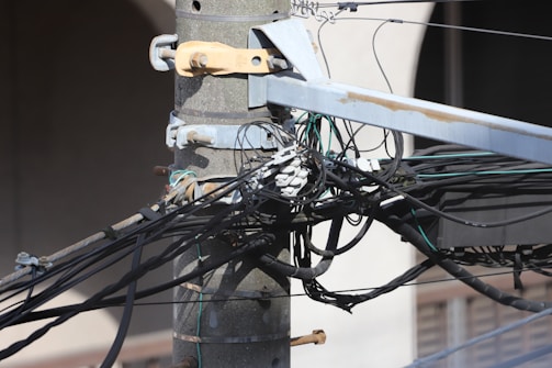 A close-up of a utility pole with numerous tangled cables and wires connected to it. The pole is made of concrete and is secured with metal brackets. There are several black and green wires along with connectors visible.
