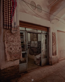 An old shop with a rustic facade featuring half-open glass doors. The entrance is adorned with hanging rugs and textiles of various patterns and colors. The dimly lit interior reveals more textiles stacked inside.