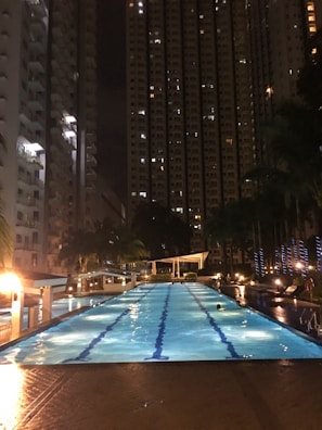 A large illuminated swimming pool bordered by tall buildings is seen at night. The pool is lined with palm trees and features ambient lighting, creating a serene and captivating atmosphere. The buildings have multiple windows with some lights on, adding to the cityscape backdrop.