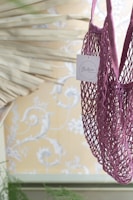 A purple netted bag with a small tag attached is in the foreground against a decorative background featuring floral patterns. The bag appears to be hanging and the tag has text on it.