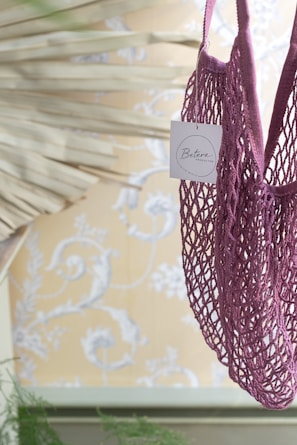 A purple netted bag with a small tag attached is in the foreground against a decorative background featuring floral patterns. The bag appears to be hanging and the tag has text on it.