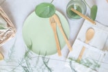 A light green ceramic plate is accompanied by eco-friendly wooden utensils, including a knife and fork. Nearby, a green glass with a plant inside sits beside a wrapped cloth napkin holding a wooden spoon. The setting features natural elements, such as green leaves and a woven basket, creating a fresh and organic atmosphere.