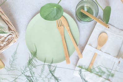 A light green ceramic plate is accompanied by eco-friendly wooden utensils, including a knife and fork. Nearby, a green glass with a plant inside sits beside a wrapped cloth napkin holding a wooden spoon. The setting features natural elements, such as green leaves and a woven basket, creating a fresh and organic atmosphere.