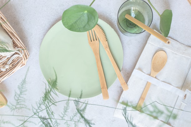 A light green ceramic plate is accompanied by eco-friendly wooden utensils, including a knife and fork. Nearby, a green glass with a plant inside sits beside a wrapped cloth napkin holding a wooden spoon. The setting features natural elements, such as green leaves and a woven basket, creating a fresh and organic atmosphere.