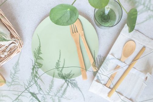 A table setting featuring pastel green tones with eco-friendly elements. A green plate is accompanied by bamboo cutlery, including a fork and knife, placed on the plate. To the right, there is a cutlery pouch holding a spoon and chopstick. A transparent green glass sits nearby, alongside decorative greenery and woven textures.