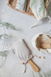 A natural soap brush with a wooden handle is placed on a light textured surface, surrounded by a wicker basket and fabric pouches adorned with fern prints. One pouch appears to be open, revealing a powdery substance inside.